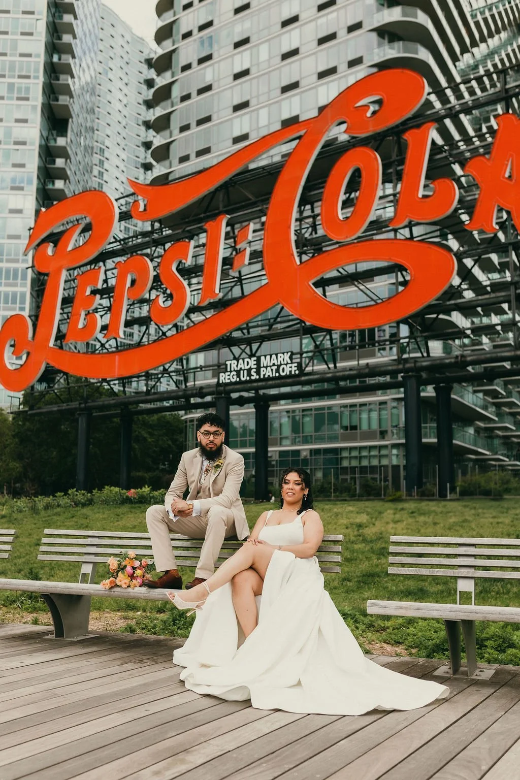A bride and groom sitting on a park bench in front of a large Pei Pei sign with modern buildings in the background. The groom is wearing a beige suit and glasses, and the bride is in a white wedding dress with one leg crossed over the other. A bouque