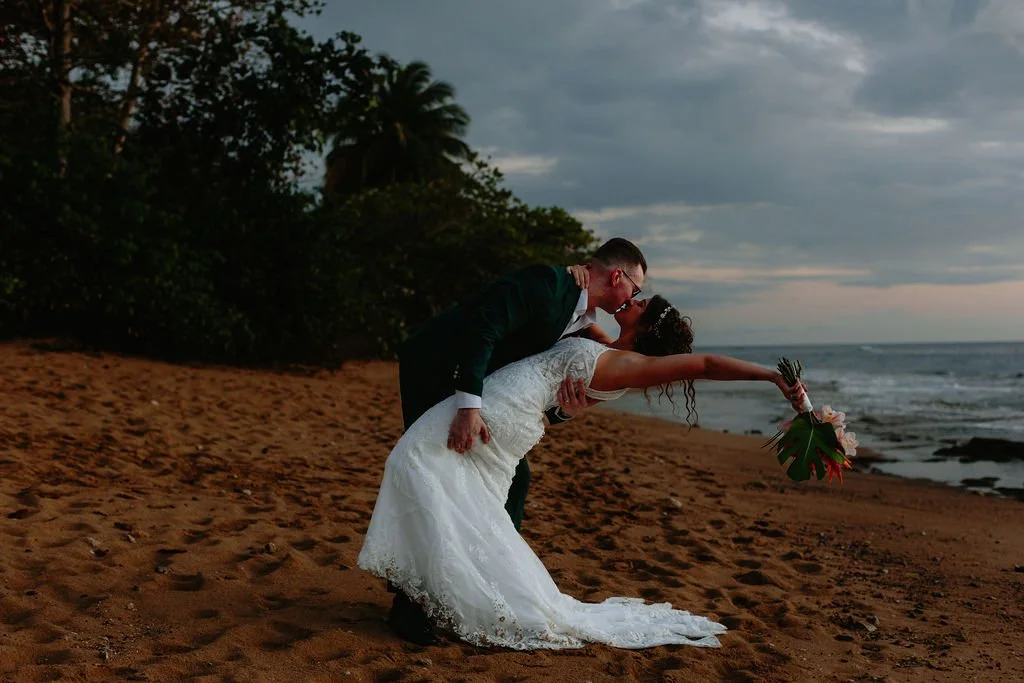 A newlywed couple sharing a kiss on the beach at sunset, with the groom dipping the bride backward while she holds a bouquet of flowers.