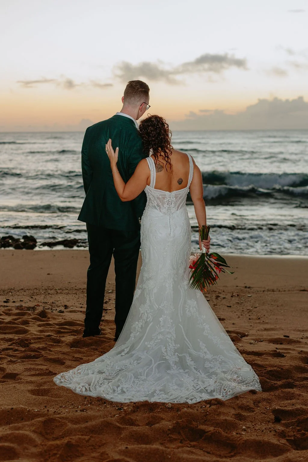 A couple dressed in wedding attire standing on a beach at sunset, with the bride holding a bouquet of flowers and the groom with his arm around her.