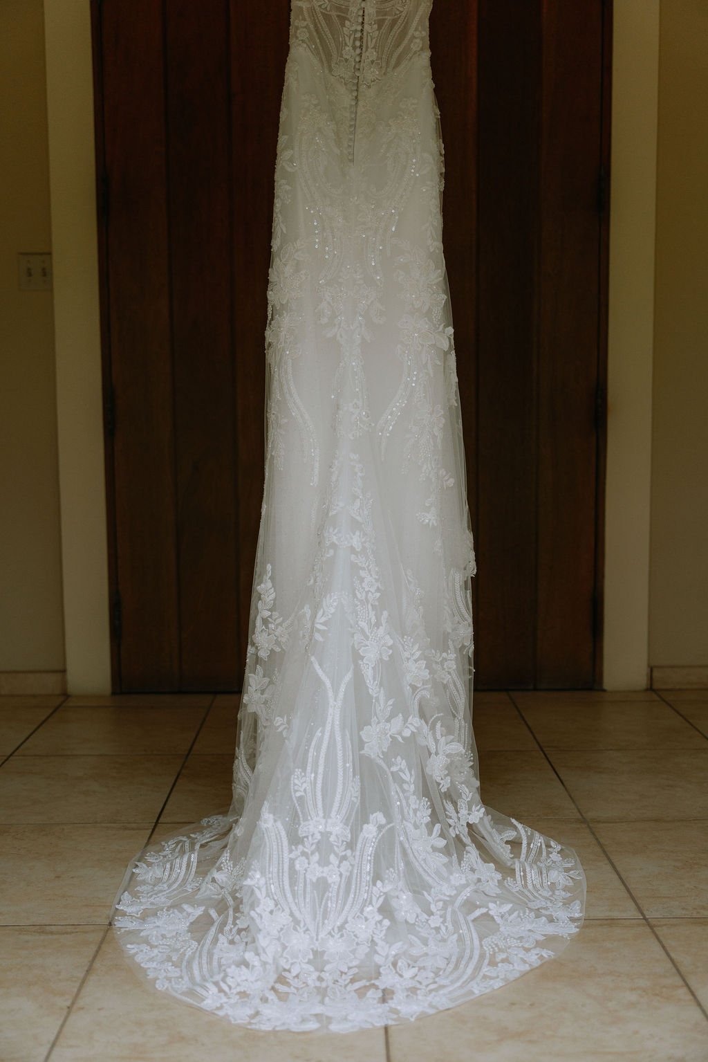 A white wedding dress hanging against a wooden door with intricate lace and embroidery details.