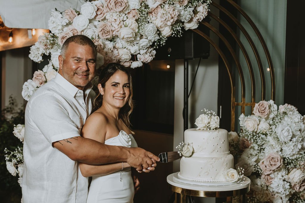 A man and a woman are smiling and cutting a white wedding cake together at a wedding reception, with a backdrop of white and blush pink flowers and a gold arch.