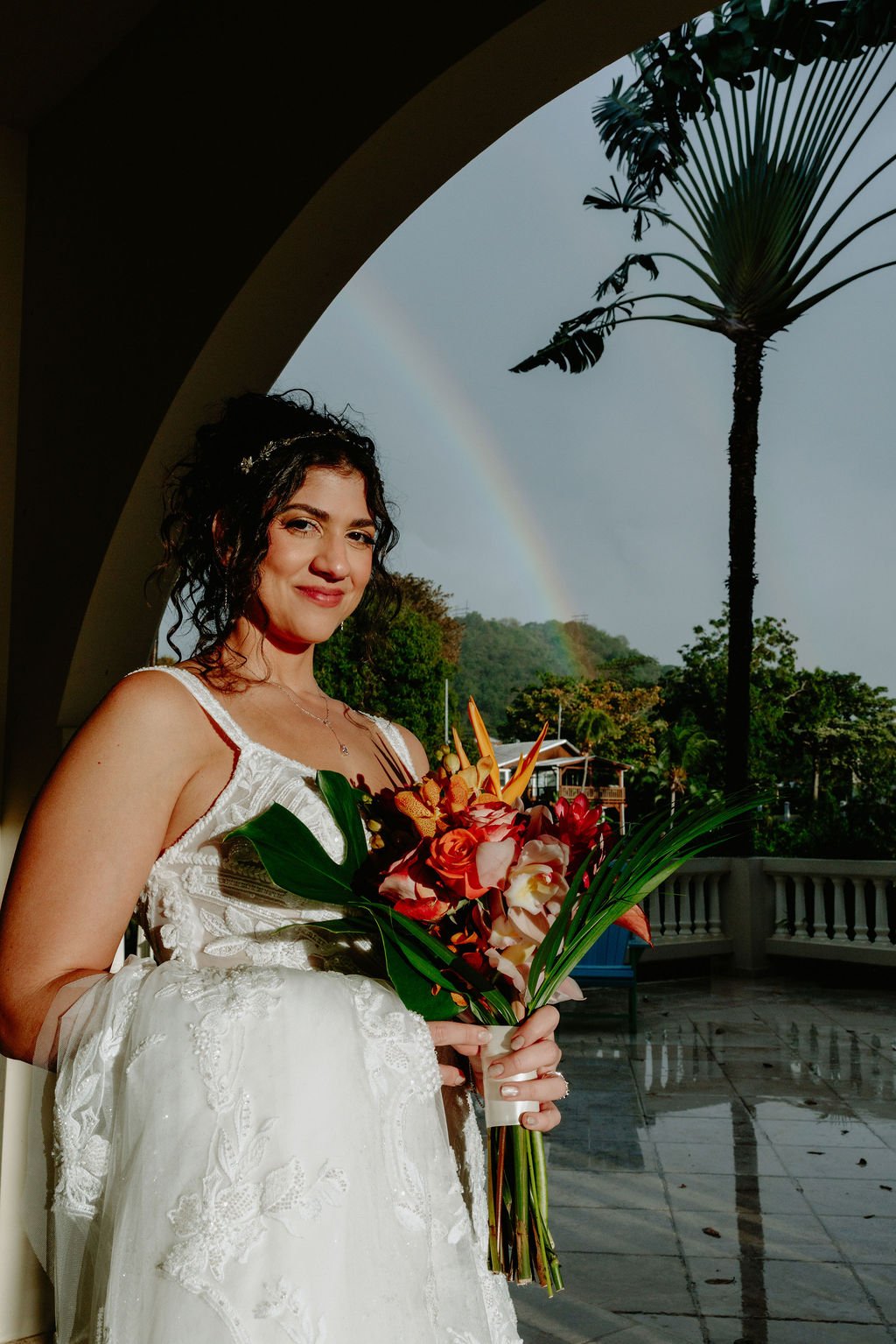 A woman in a white dress holding a bouquet of flowers, standing under an archway with a rainbow and lush greenery in the background.