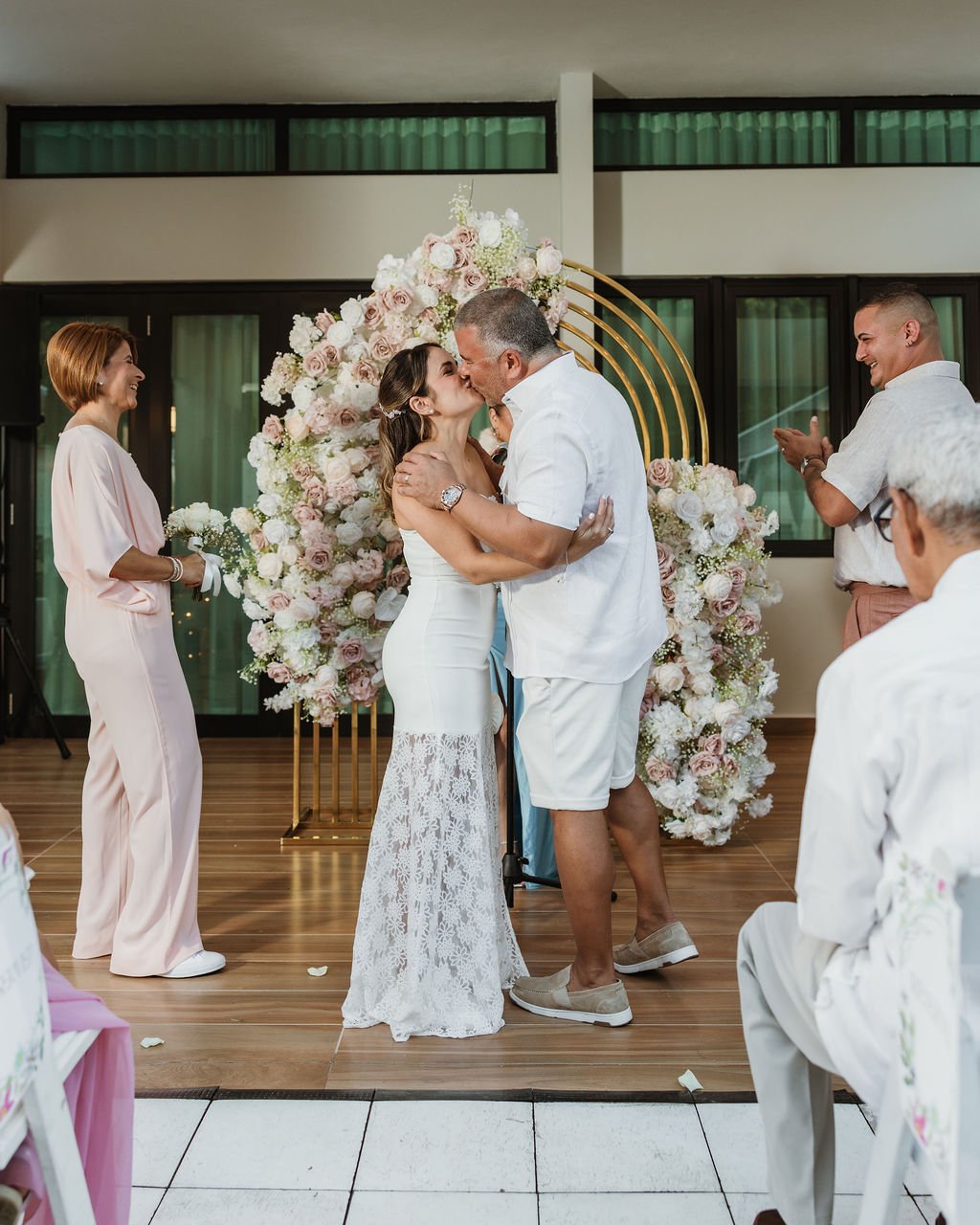 A couple kisses during a wedding ceremony with a floral backdrop, while guests celebrate and look on happily.