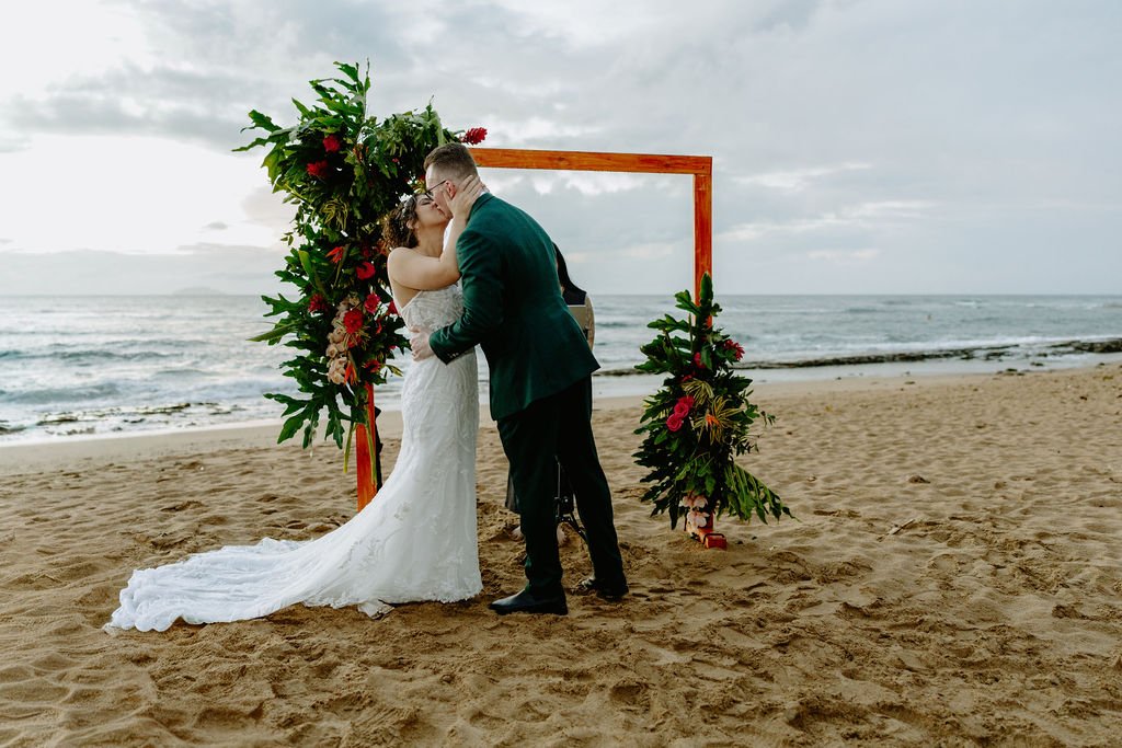 A couple getting married and sharing a kiss on the beach during their wedding ceremony, with an ocean view and cloudy sky in the background, framed by a floral arch.