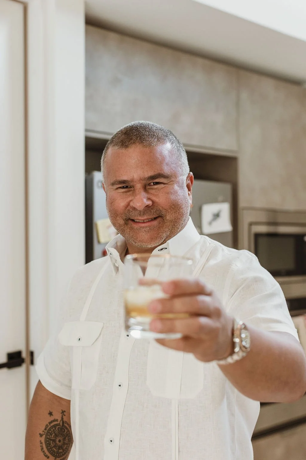 A man with short hair and a white shirt smiling and holding a glass of beverage toward the camera in a kitchen.