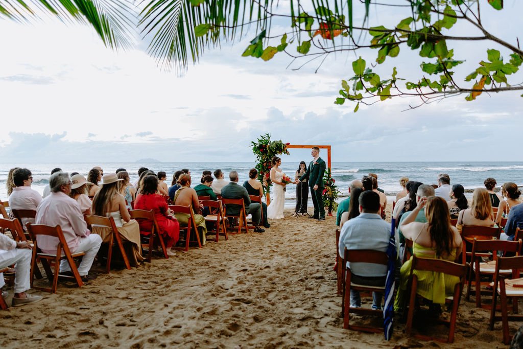 Beach wedding ceremony with guests seated in chairs, facing an officiant and a couple exchanging vows under a floral arch, with the ocean and cloudy sky in the background.