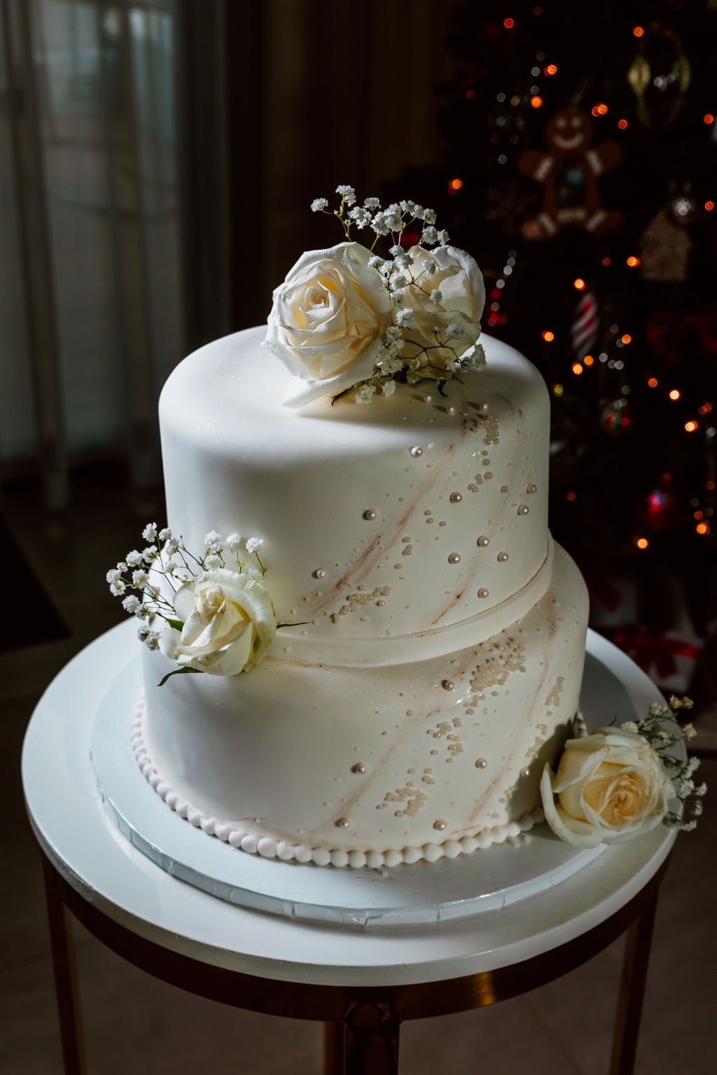 A three-tiered white wedding cake decorated with white roses, baby's breath, and small silver beads, placed on a white cake stand.