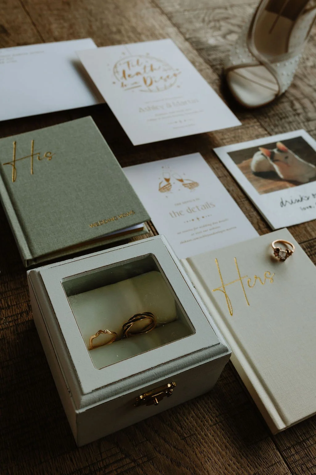 Wedding rings in a jewelry box on a wooden table surrounded by wedding invitation cards, a guest book, and a photo of a duck, with a glass of drink and a high-heeled shoe in the background.