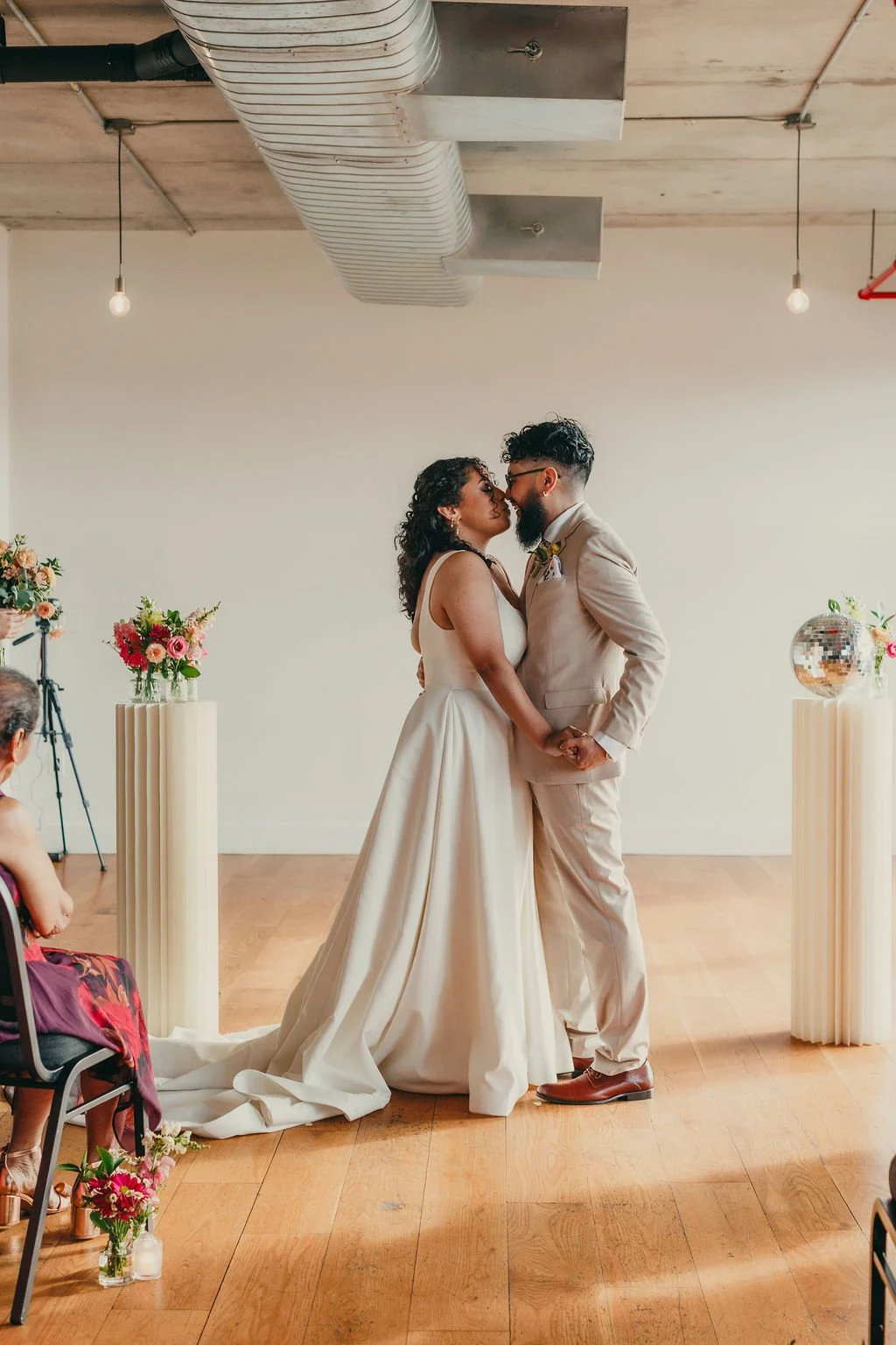 A couple kissing at their wedding ceremony in a modern, minimalist venue with wooden floors and white walls, decorated with flower arrangements on tall pedestals.