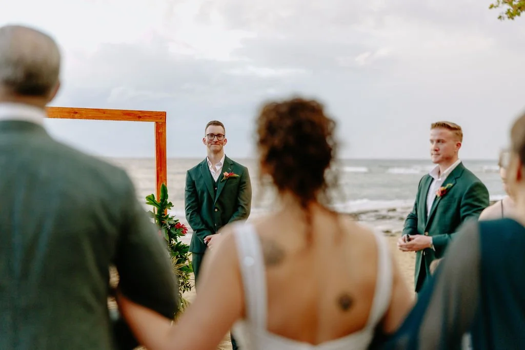 A couple getting married on the beach, standing under a wooden arch decorated with flowers, with guests witnessing the ceremony.