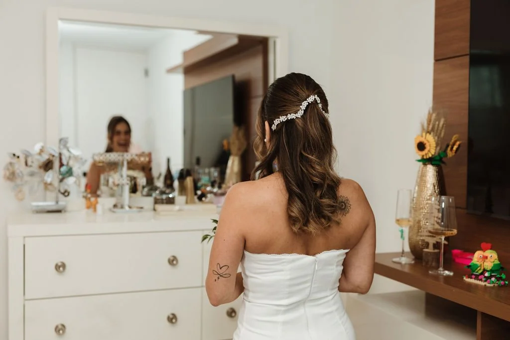 A woman in a white strapless dress with a decorative headband looks at her reflection in a mirror. The room is decorated with flowers and drinks, possibly preparing for a special occasion.