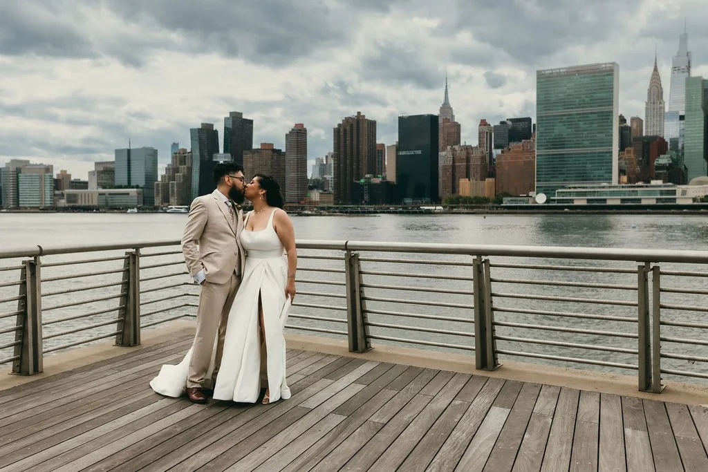 A couple stands on a waterfront promenade with New York City skyline in the background, sharing a kiss.