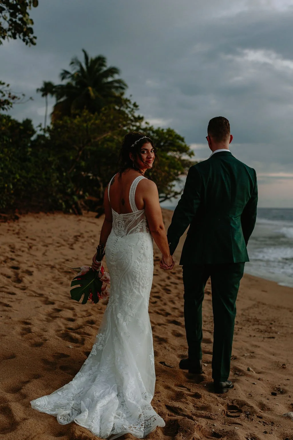 A bride in a white lace wedding dress and a groom in a dark suit walk hand-in-hand along a sandy beach at sunset, with the bride looking back and smiling toward the camera.