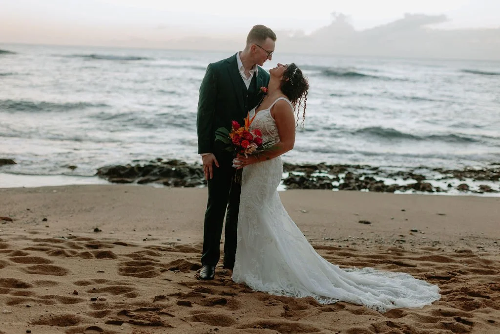 A newlywed couple on the beach, standing close together, with the ocean in the background. The bride wears a white lace wedding dress and holds a colorful bouquet, and the groom wears a dark suit with glasses. They are smiling and looking into each o