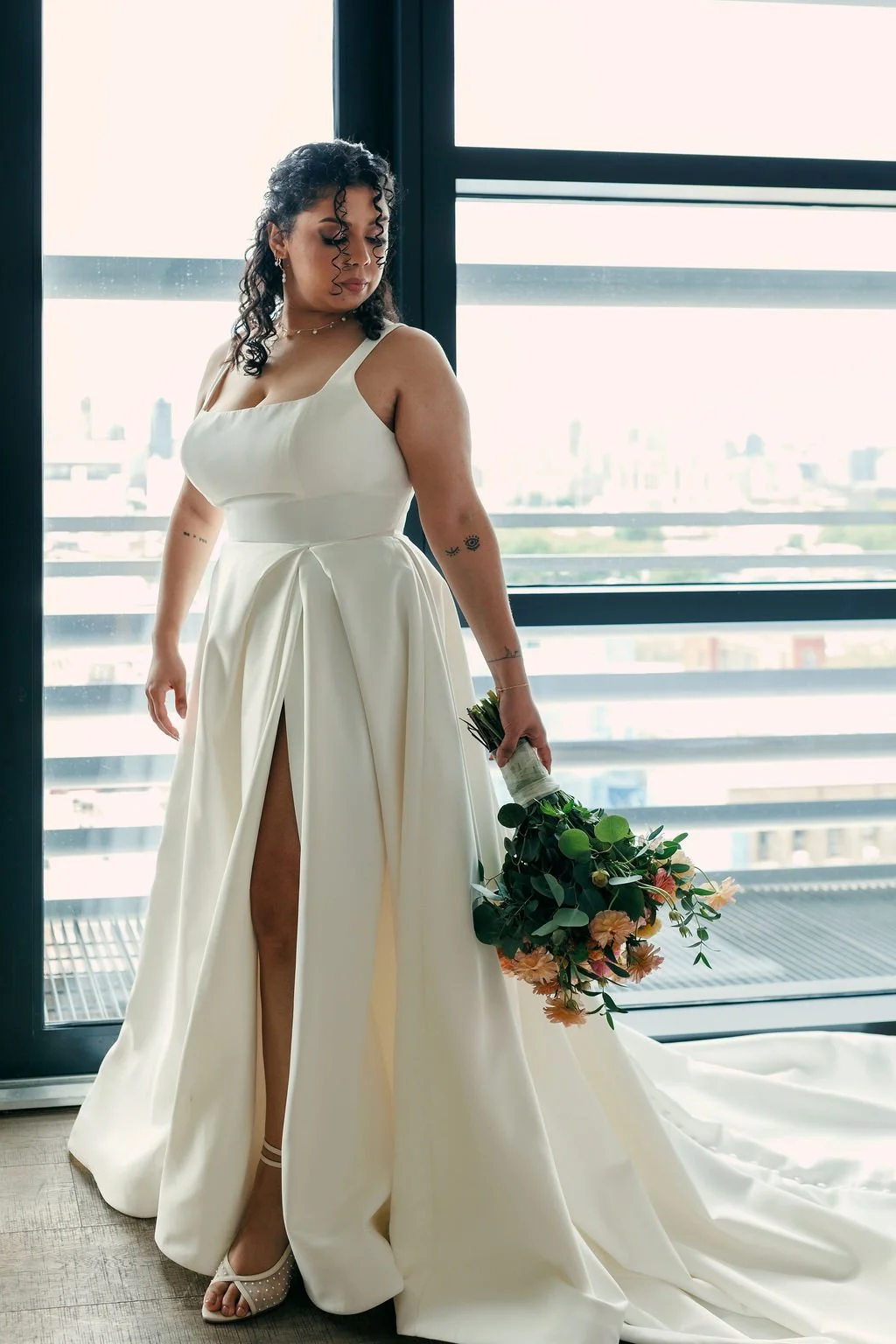 A woman in a white wedding dress holding a bouquet, standing by a large window in a bright room.