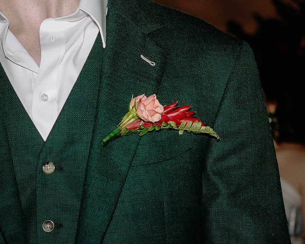 Close-up of a man's green blazer with a pink and red boutonniere on the lapel, over a white dress shirt.