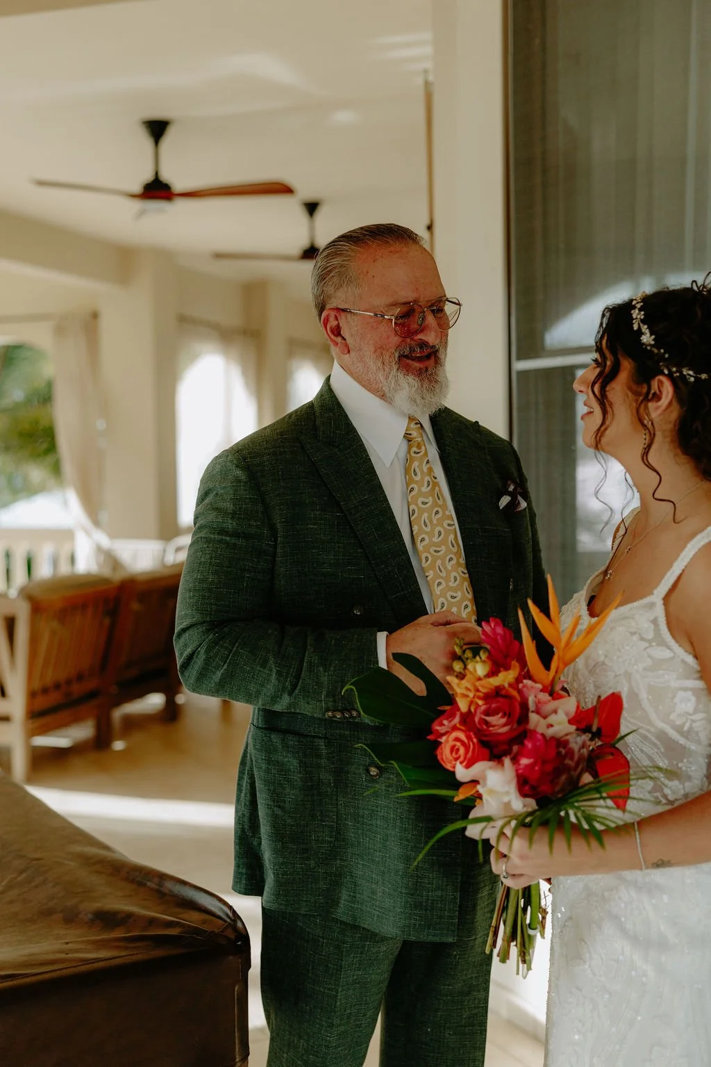 An older man in a patterned yellow tie and plaid green suit is smiling and talking to a young woman in a white wedding dress holding a bouquet of red and orange flowers in a bright, elegant room.