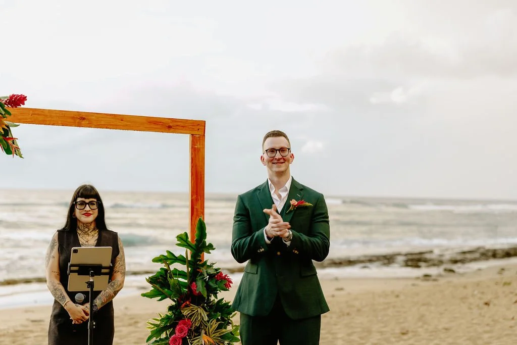A groom waiting at an outdoor beach wedding ceremony, standing in front of a wooden arch decorated with green leaves and red flowers, with an officiant woman holding a tablet and microphone nearby.