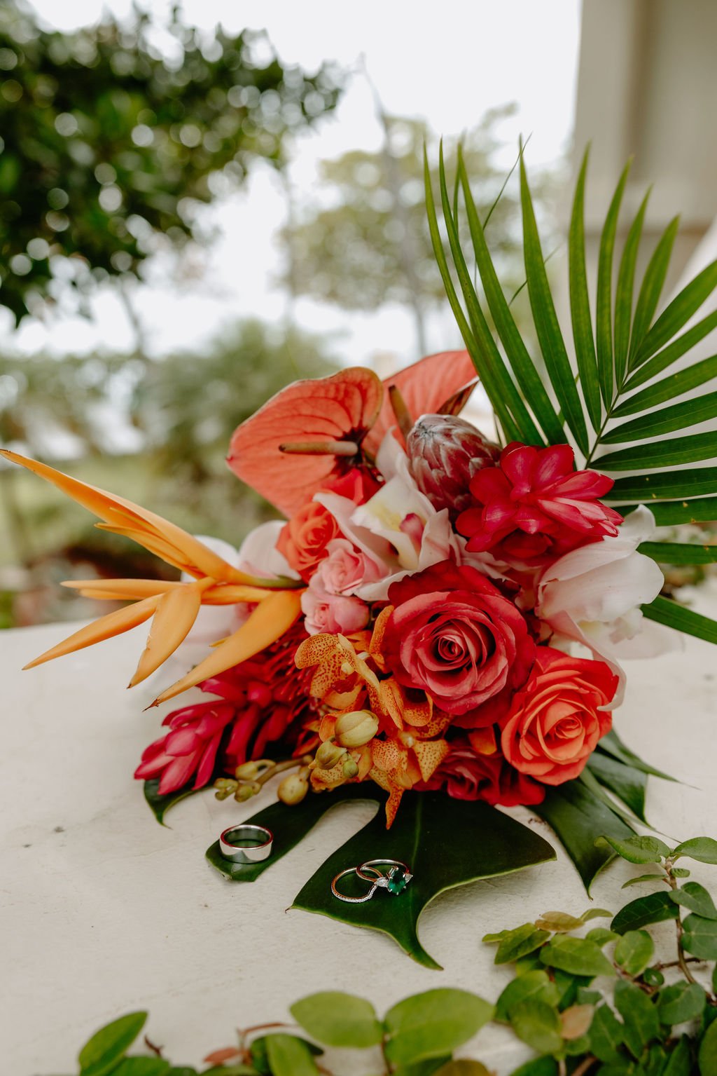 Vibrant tropical flower bouquet with orange, red, pink, and white flowers, green palm leaves, and two rings placed on a white surface.