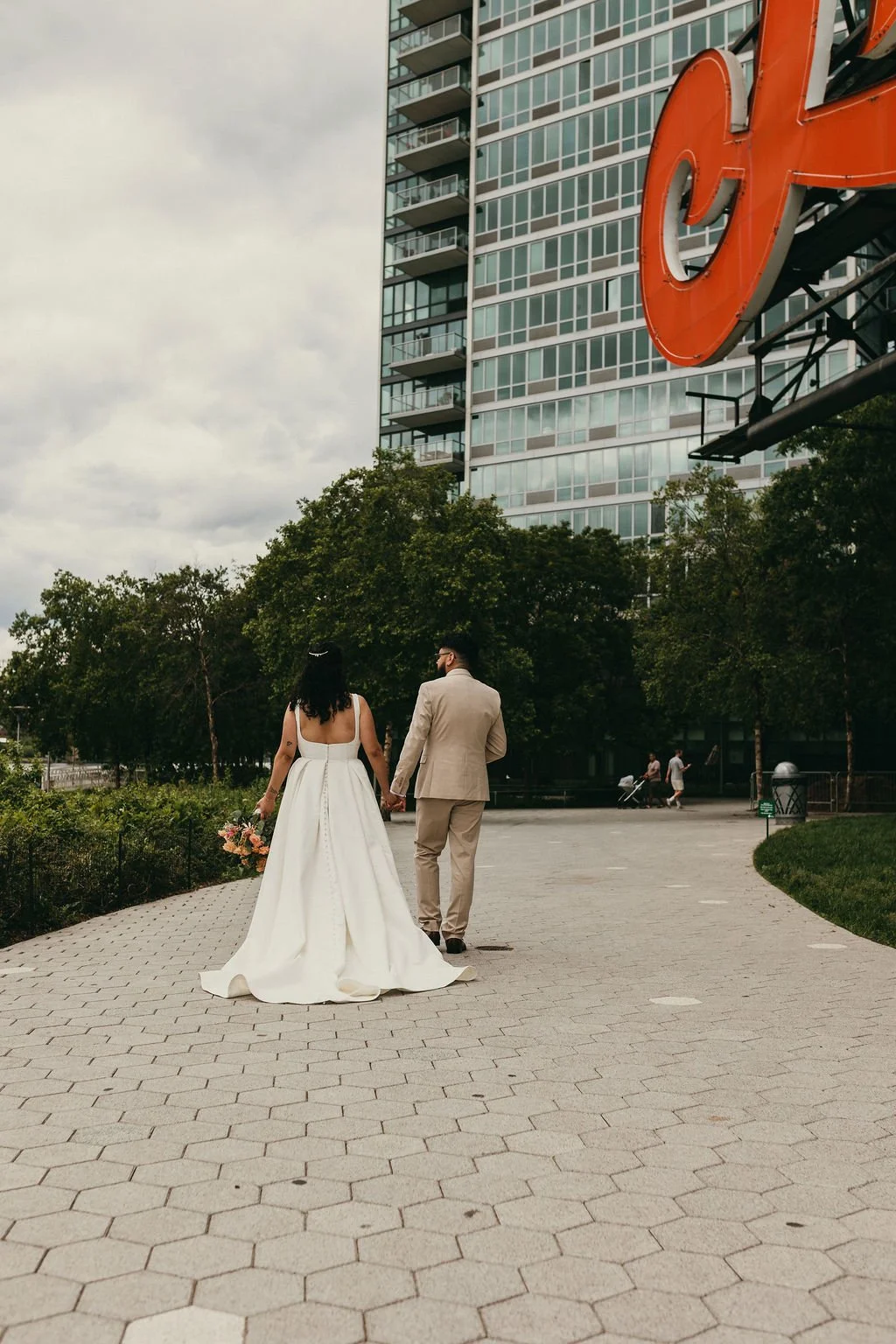 A bride in a white wedding dress and a groom in a beige suit are walking hand in hand in an urban park with green trees, in front of a tall modern building. The bride is holding a bouquet of flowers and they are walking away from the camera. There ar