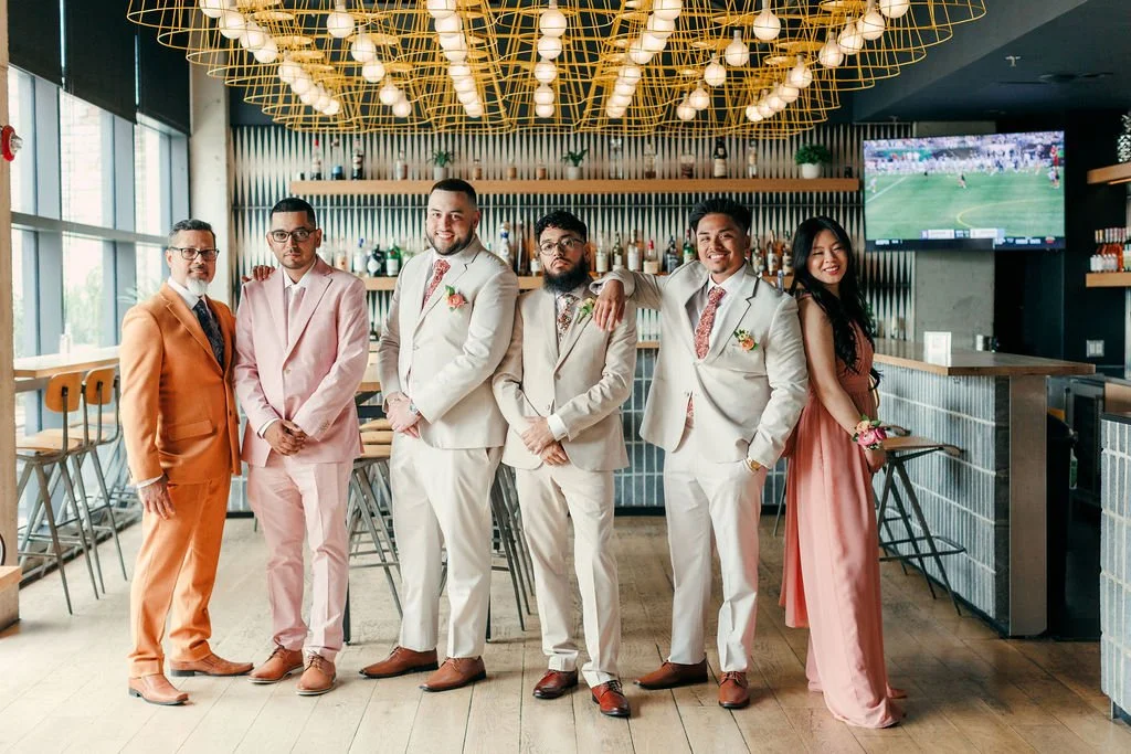 Six people in formal attire standing inside a modern restaurant. Four men are wearing pastel-colored suits, and one woman is wearing a pink dress. They are posing for a photo, smiling, with a bar, large windows, and a TV showing a soccer game in the 