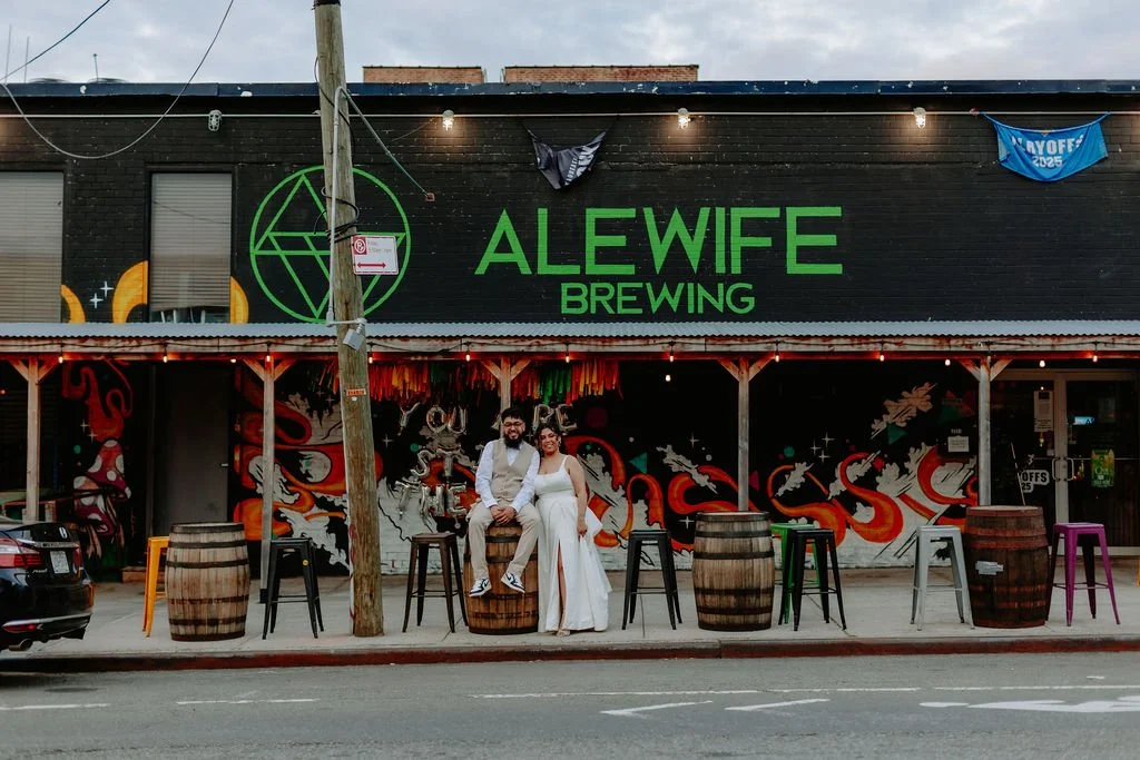 A couple dressed in wedding attire standing outside Alewife Brewing, a bar with colorful murals and barrel decorations on the sidewalk.