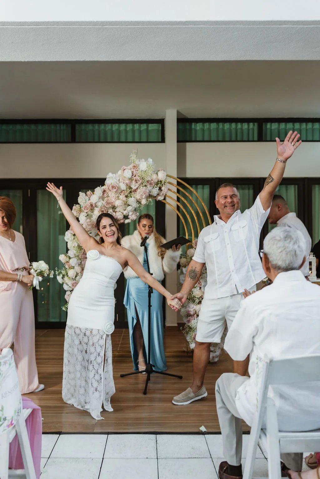 A couple holding hands and smiling during a wedding ceremony, with an officiant reading in the background, floral decorations, and guests seated around.