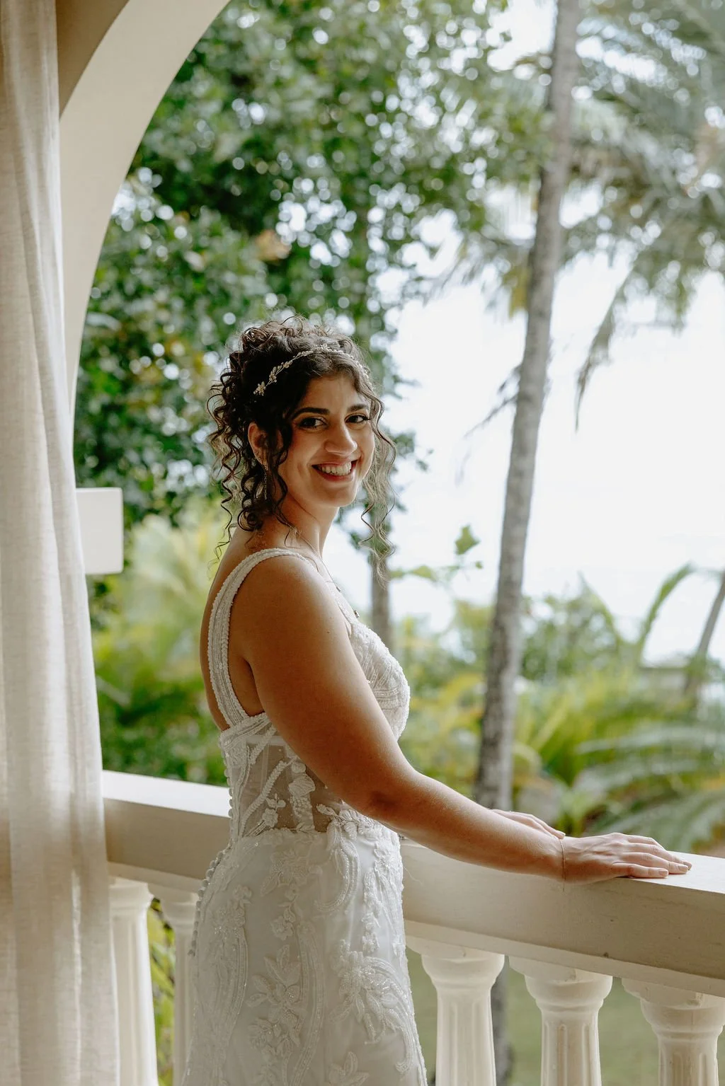 A woman in a white lace wedding dress smiling and leaning on a balcony railing with a tropical background of trees and greenery.