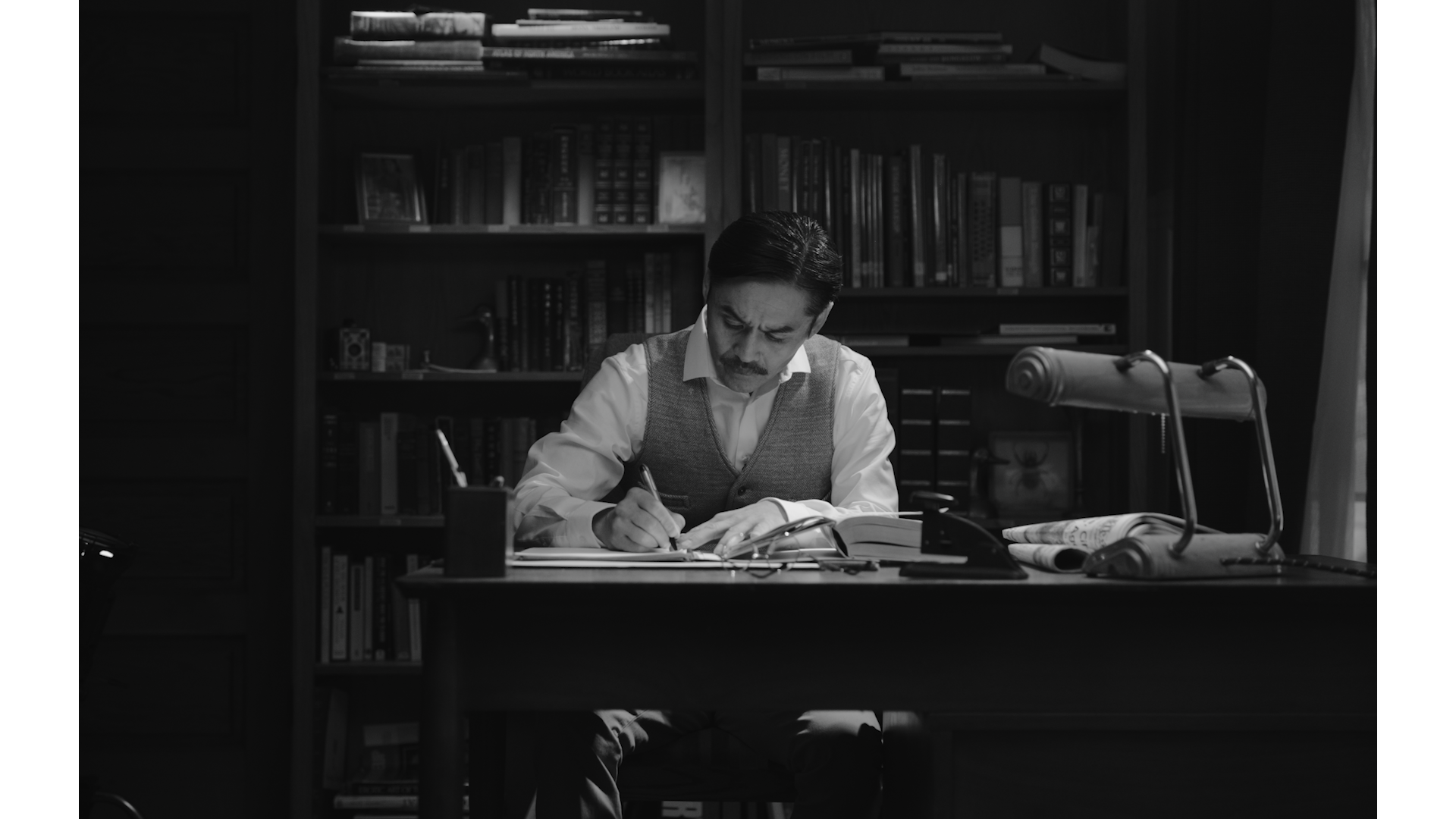 An Issei father writes at his desk, with a wall of books behind him.