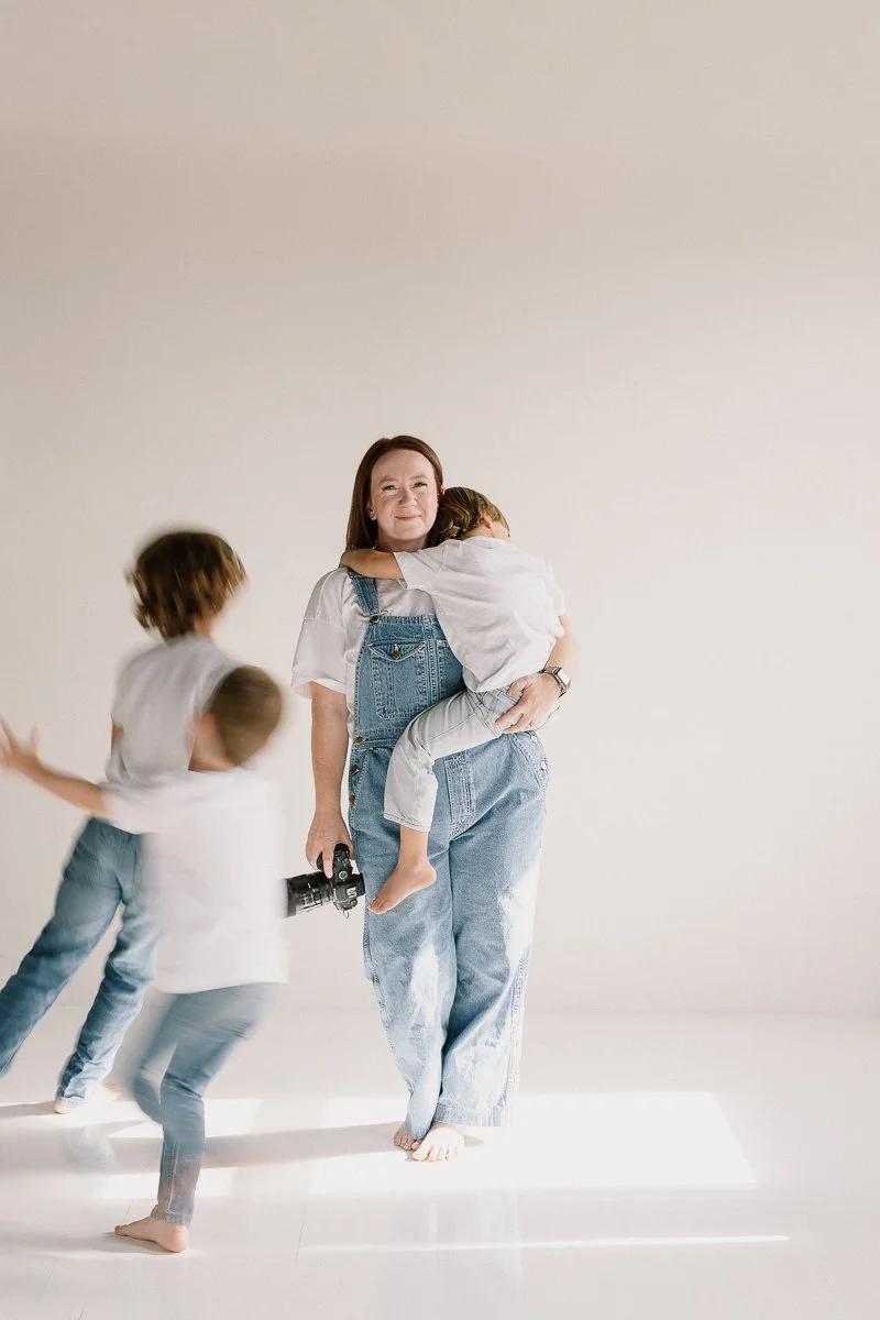 Arizona birth and family photographer holding toddler and camera while older children run around her in neutral studio space