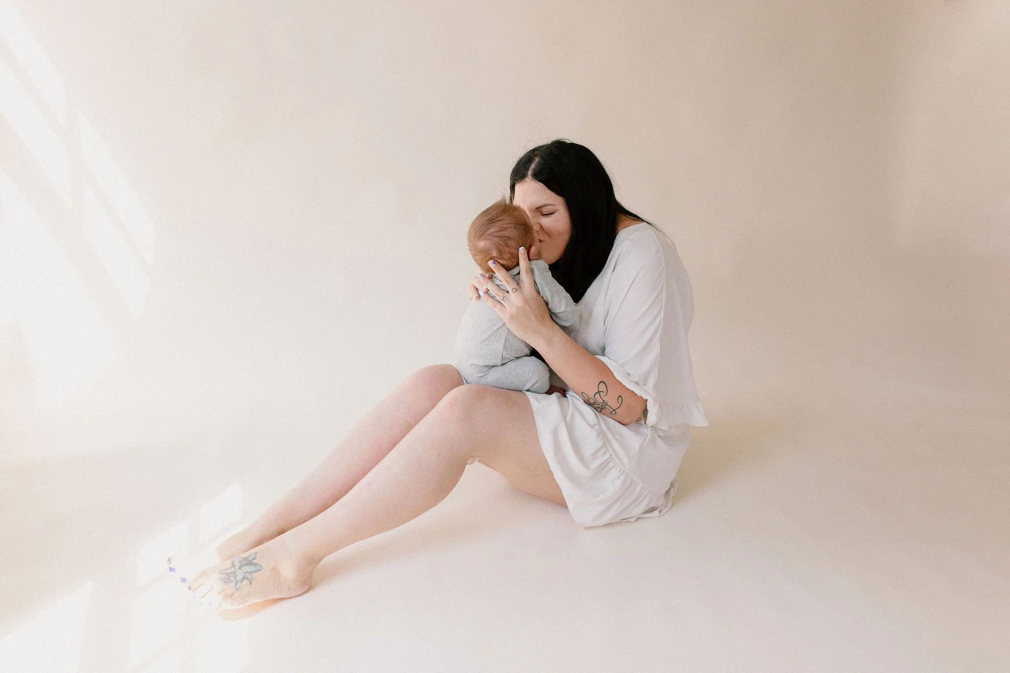 mom sitting on the floor kissing baby in neutral natural light studio by Phoenix newborn photographer