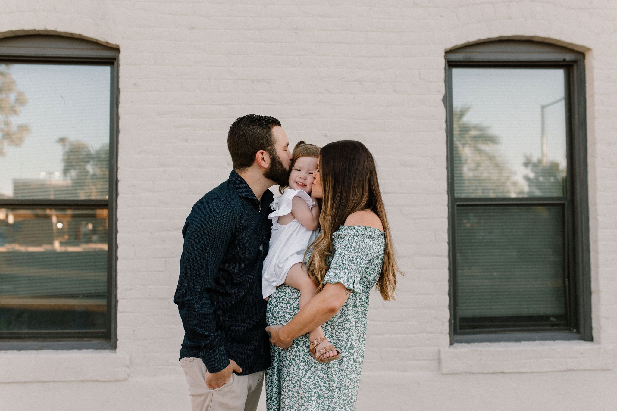 pregnant mom holding belly with toddler sitting mom and dad kissing her cheek in Gilbert Arizona maternity photography session