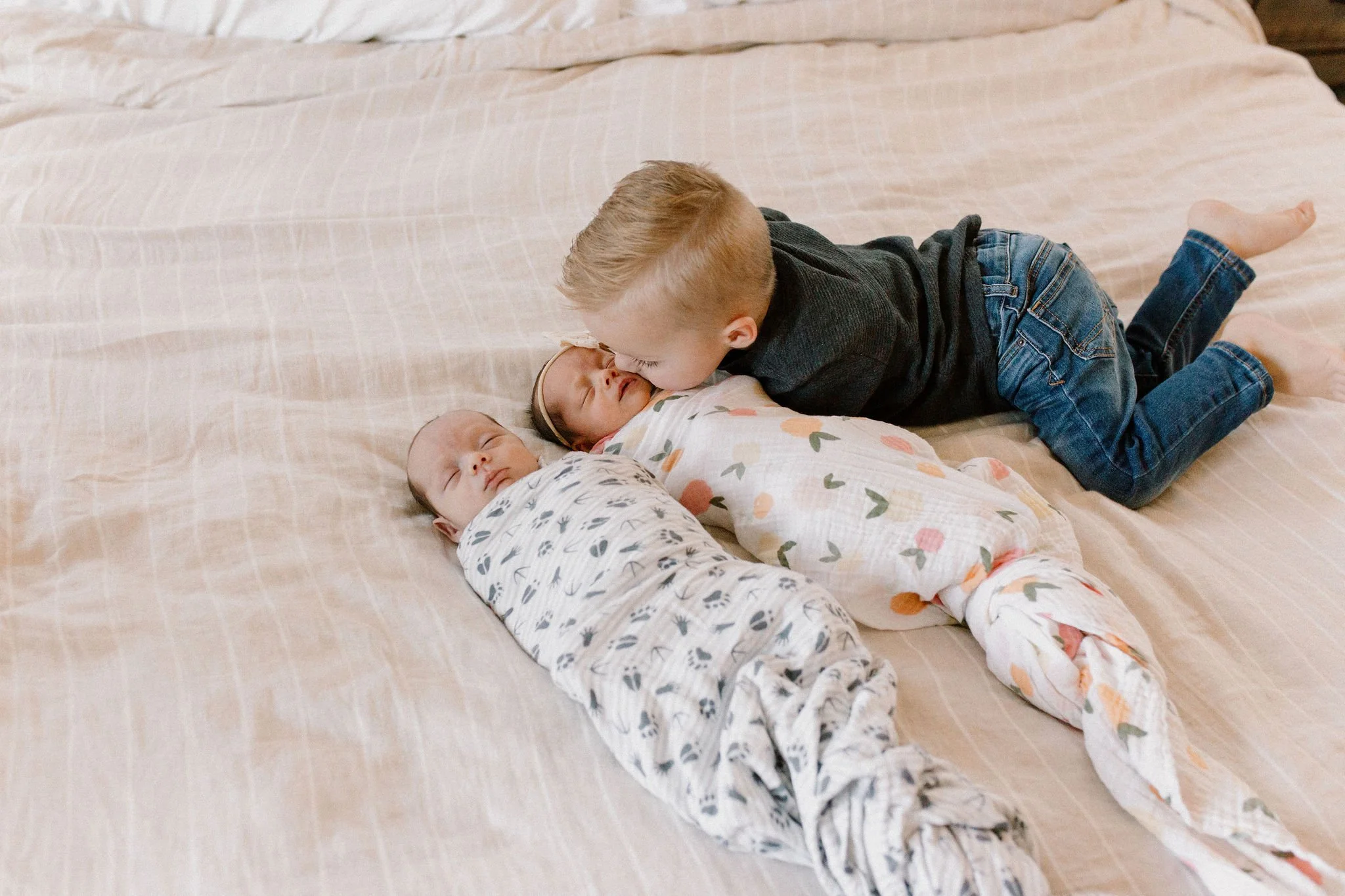 twin newborn boy and girl swaddled on bed with toddler big brother giving kisses