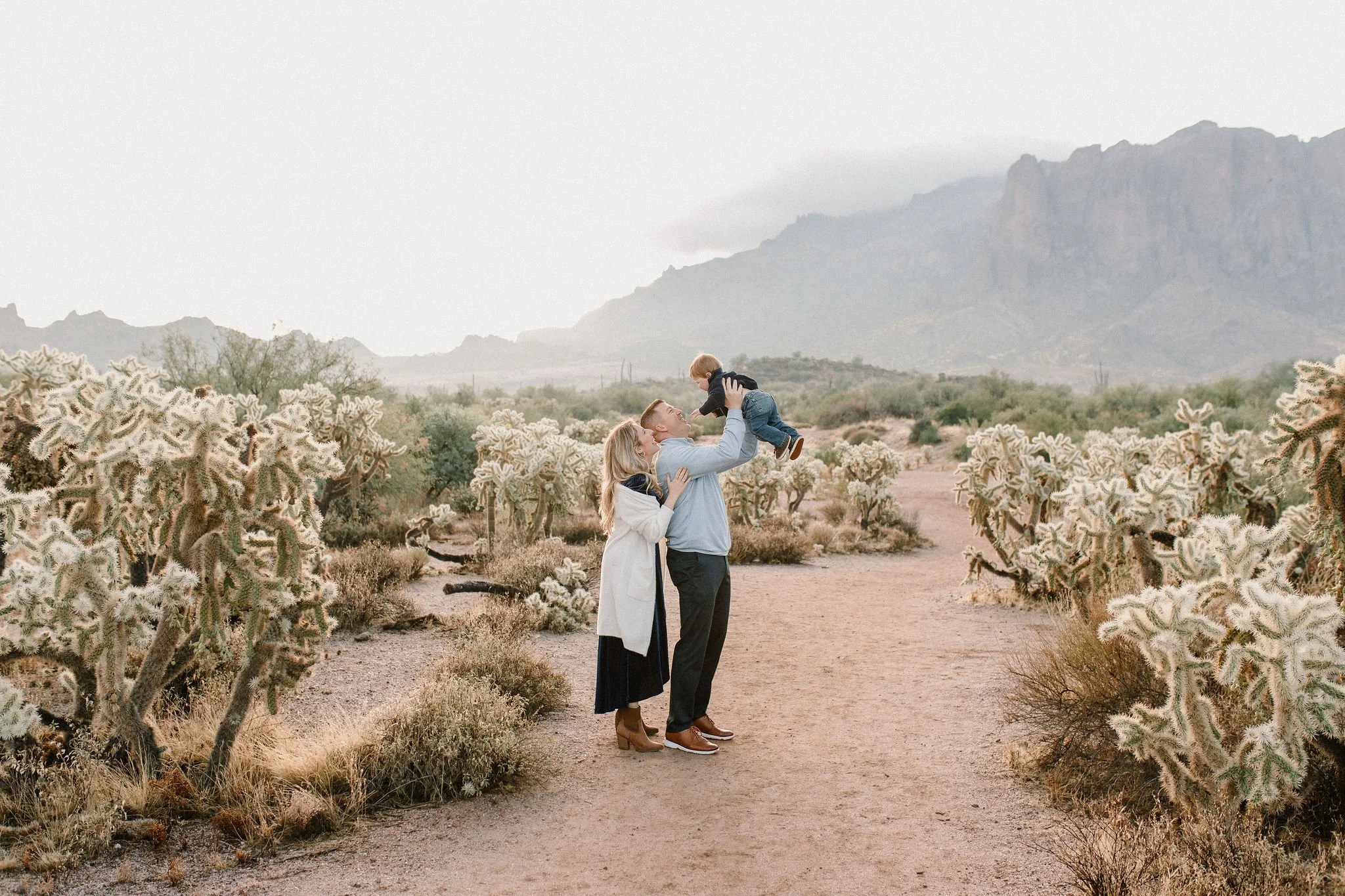 dad holding baby boy in the air with mom looking at them in desert misty morning photography session at the Superstition Mountains