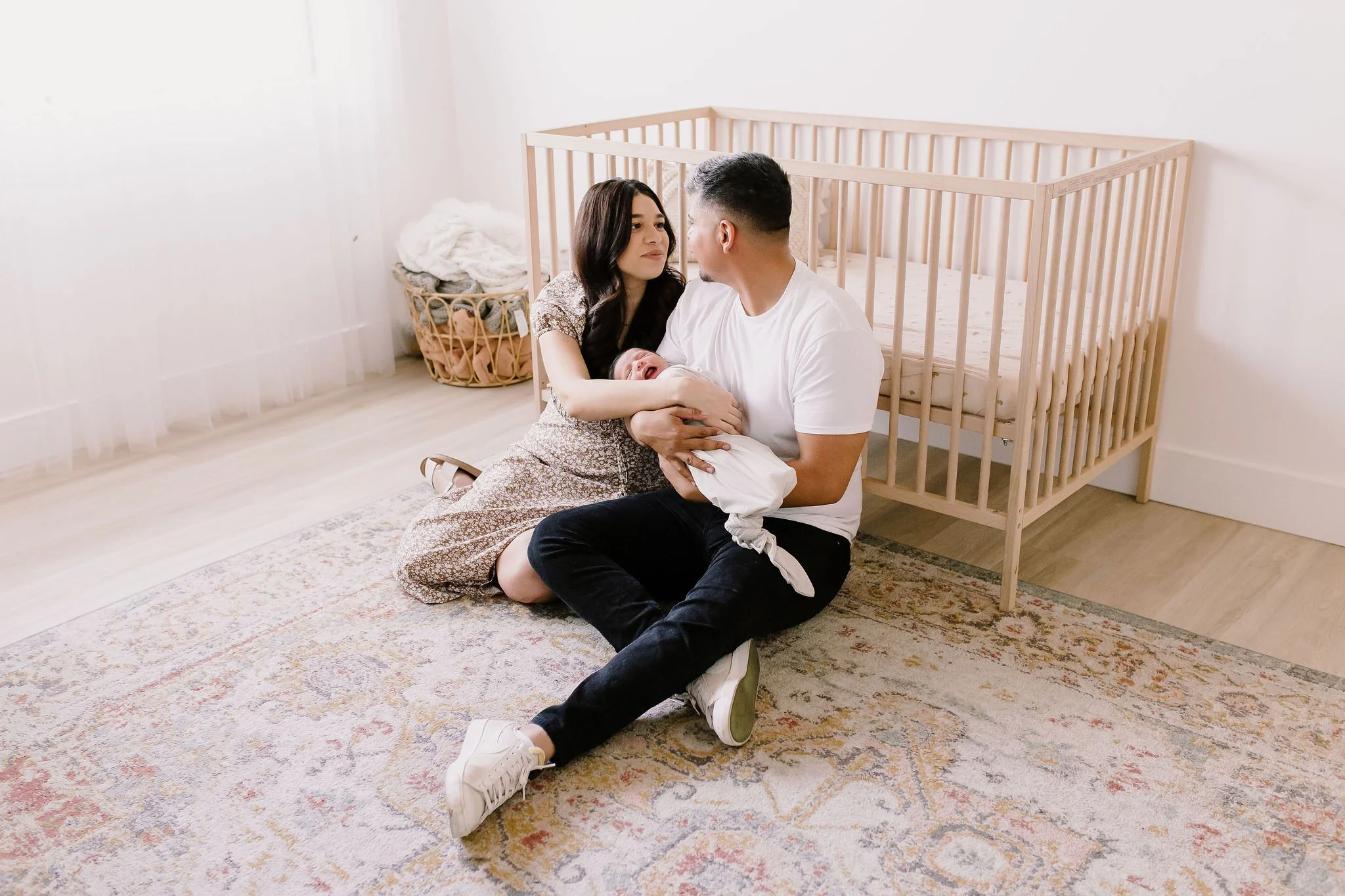 mom and dad sitting on the floor in the nursery in front of crib holding baby in studio newborn photography session