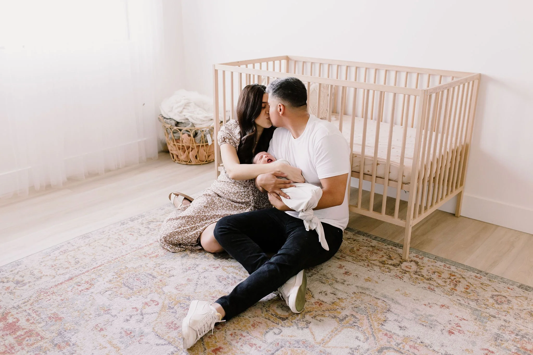 mom and dad kissing and sitting on the floor in the nursery in front of crib holding baby in studio newborn photography session