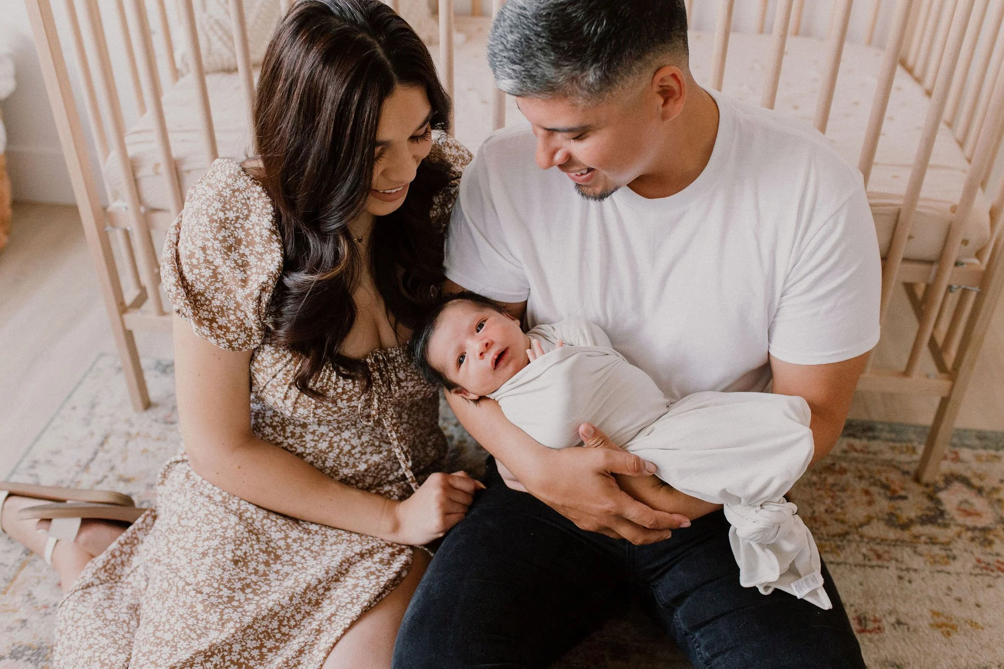 mom and dad sitting on the nursery floor in front of crib holding baby in studio newborn photography session