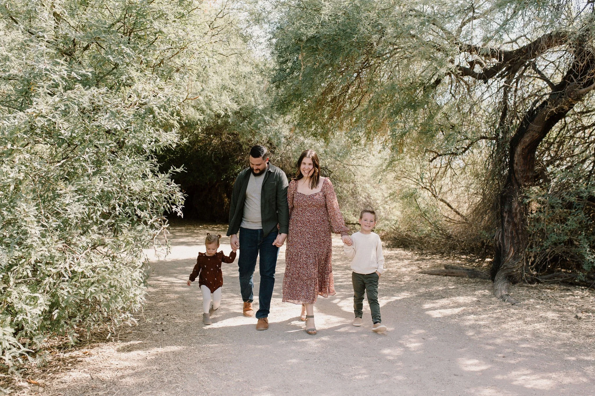 Family holding hands walking in green desert setting while smiling at Gilbert Arizona family photographer