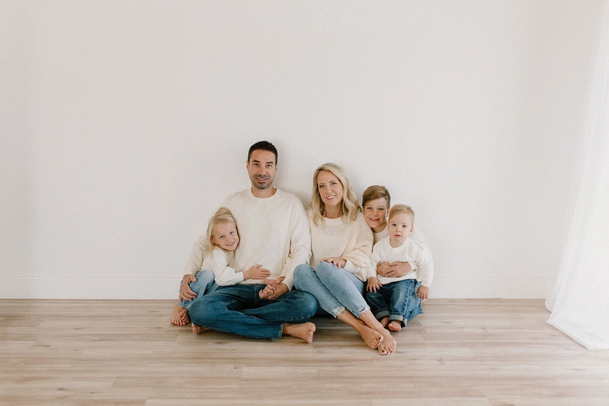 family wearing white sweaters and blue jeans sitting together in neutral studio space