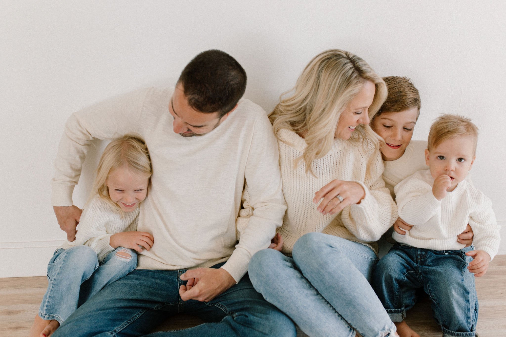 family of five sitting together in white sweaters and blue jeans cuddling and tickling in a neutral studio pace