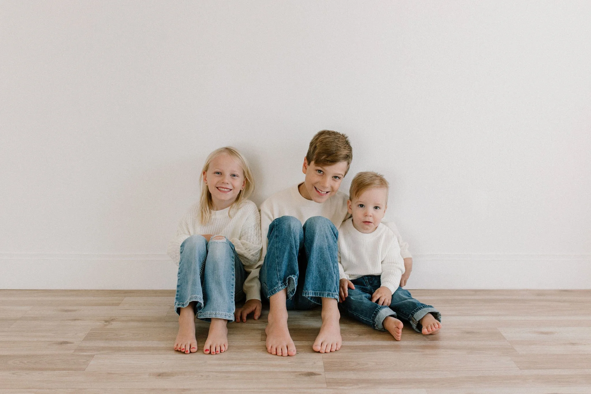 siblings wearing white sweaters and blue jeans sitting together in neutral studio space