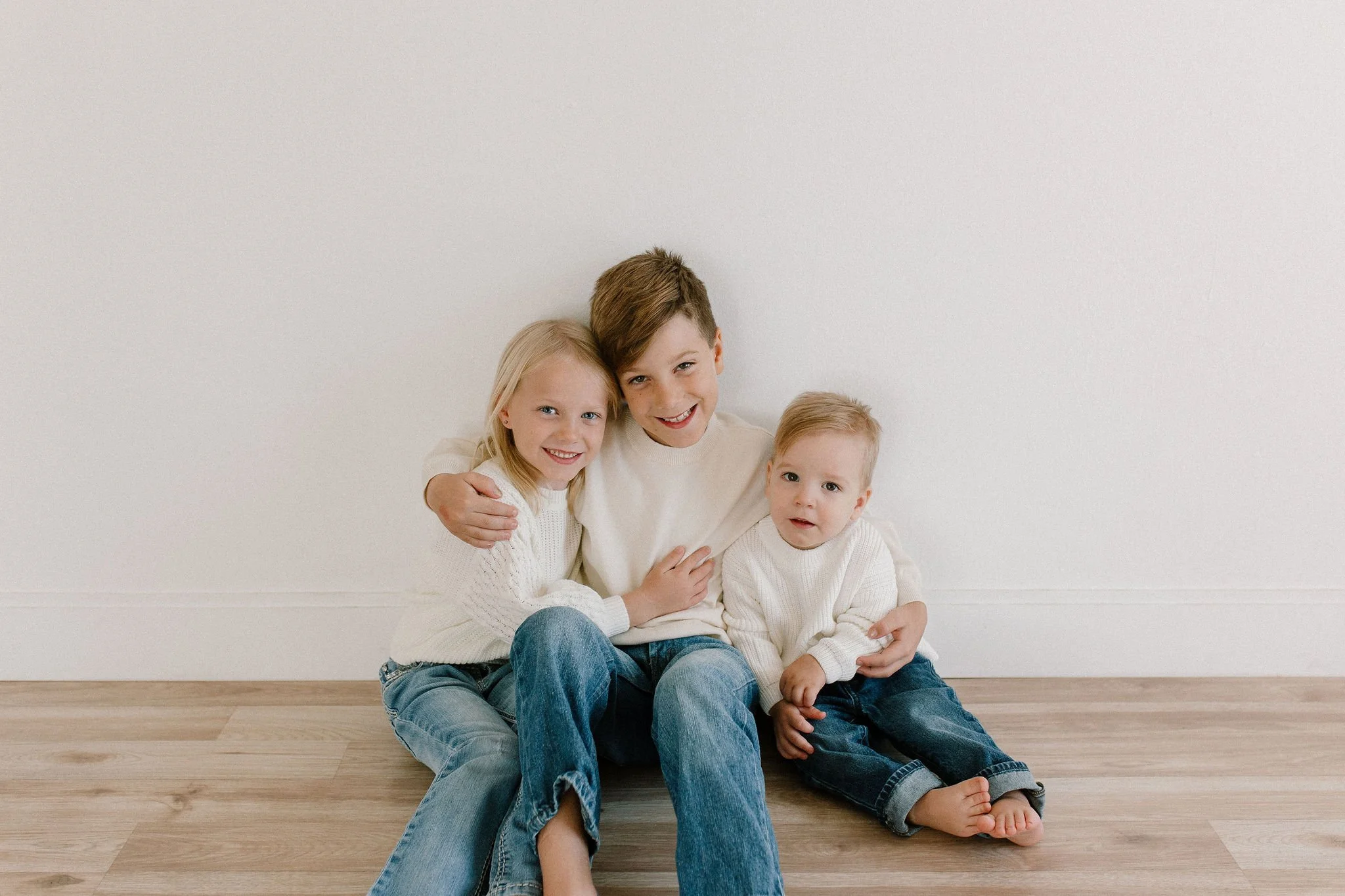 siblings wearing white sweaters and blue jeans sitting together in neutral studio space