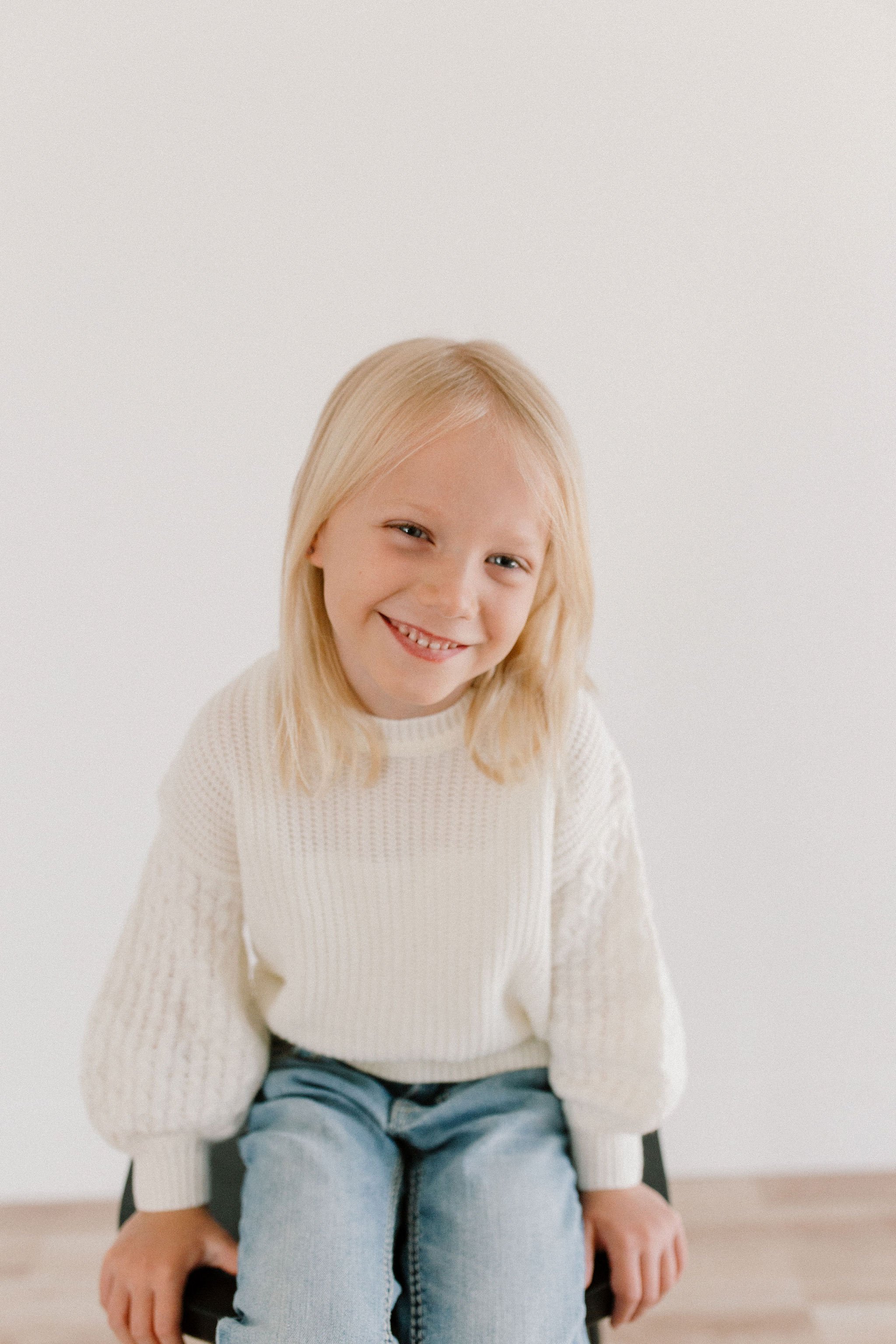 childhood portrait girl in white sweater and blue jeans sitting on a stool in neutral studio space