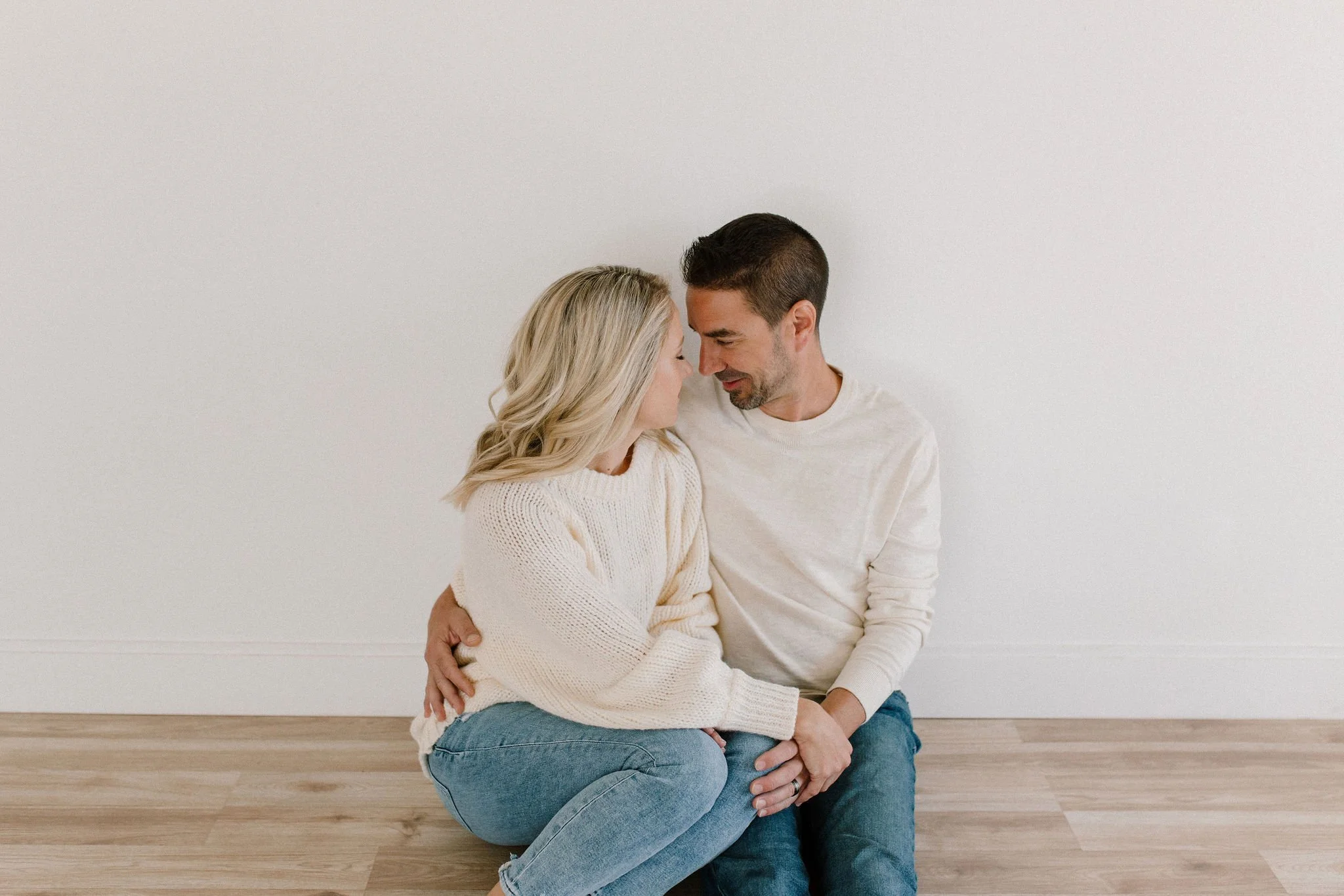 mom and dad wearing white sweaters and blue jeans sitting and cuddling in a neutral studio space