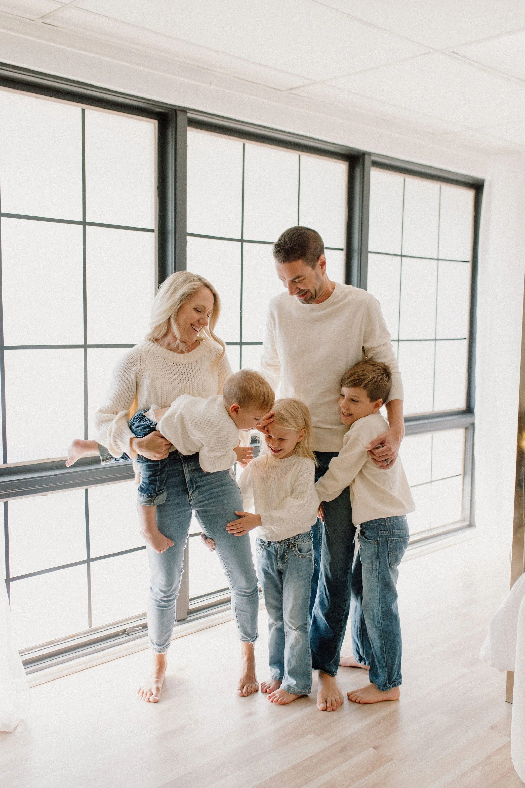 Family of five wearing white sweaters and blue jeans  standing together and playing by a window in a neutral studio