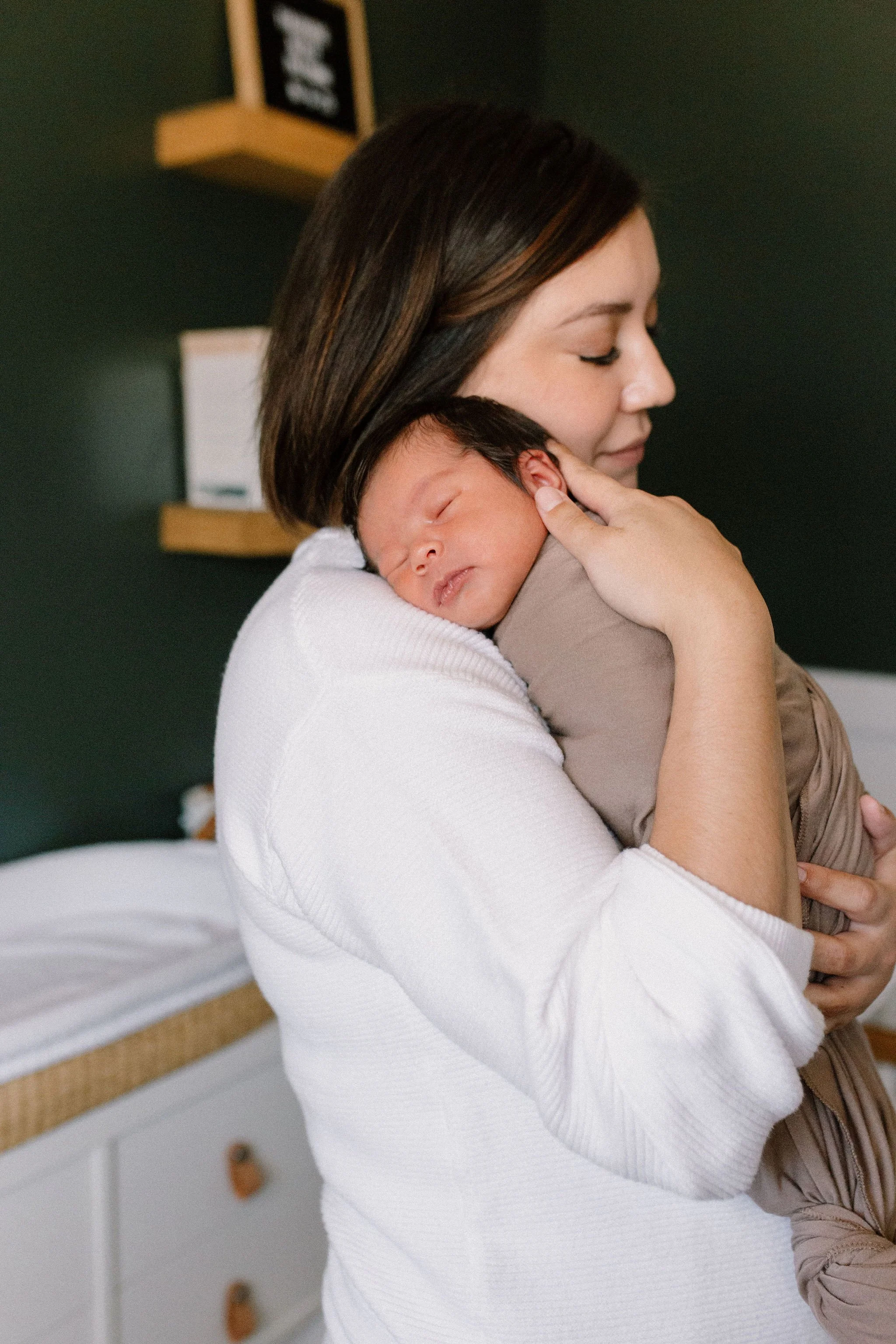 swaddled newborn baby laying on mom's shoulder in dark green nursery