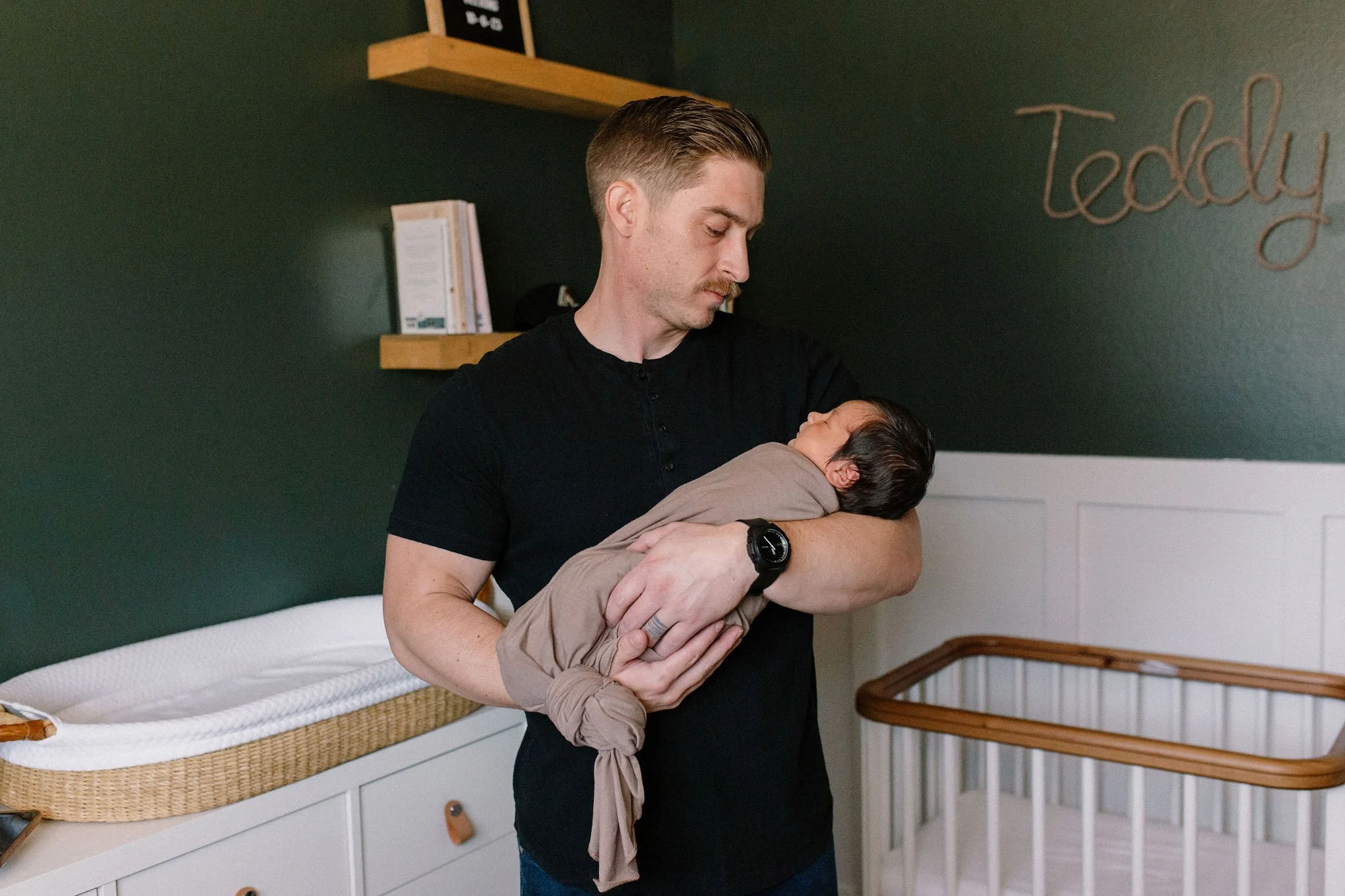 dad holding swaddled newborn baby in dark green nursery