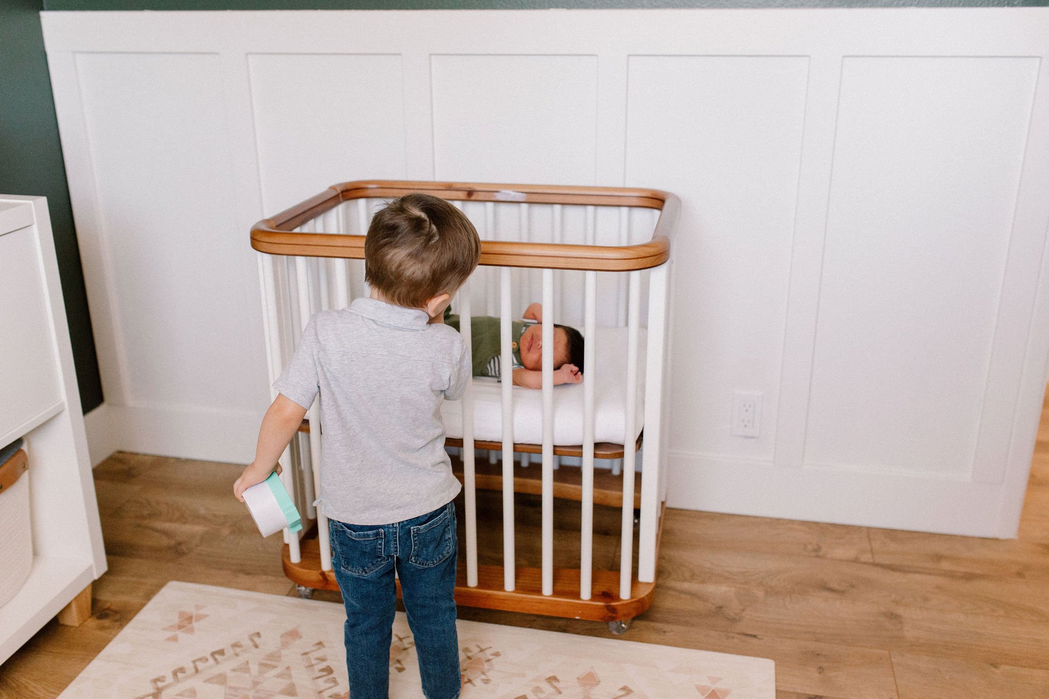 older brother looking at sleeping newborn baby in bassinet in dark green nursery