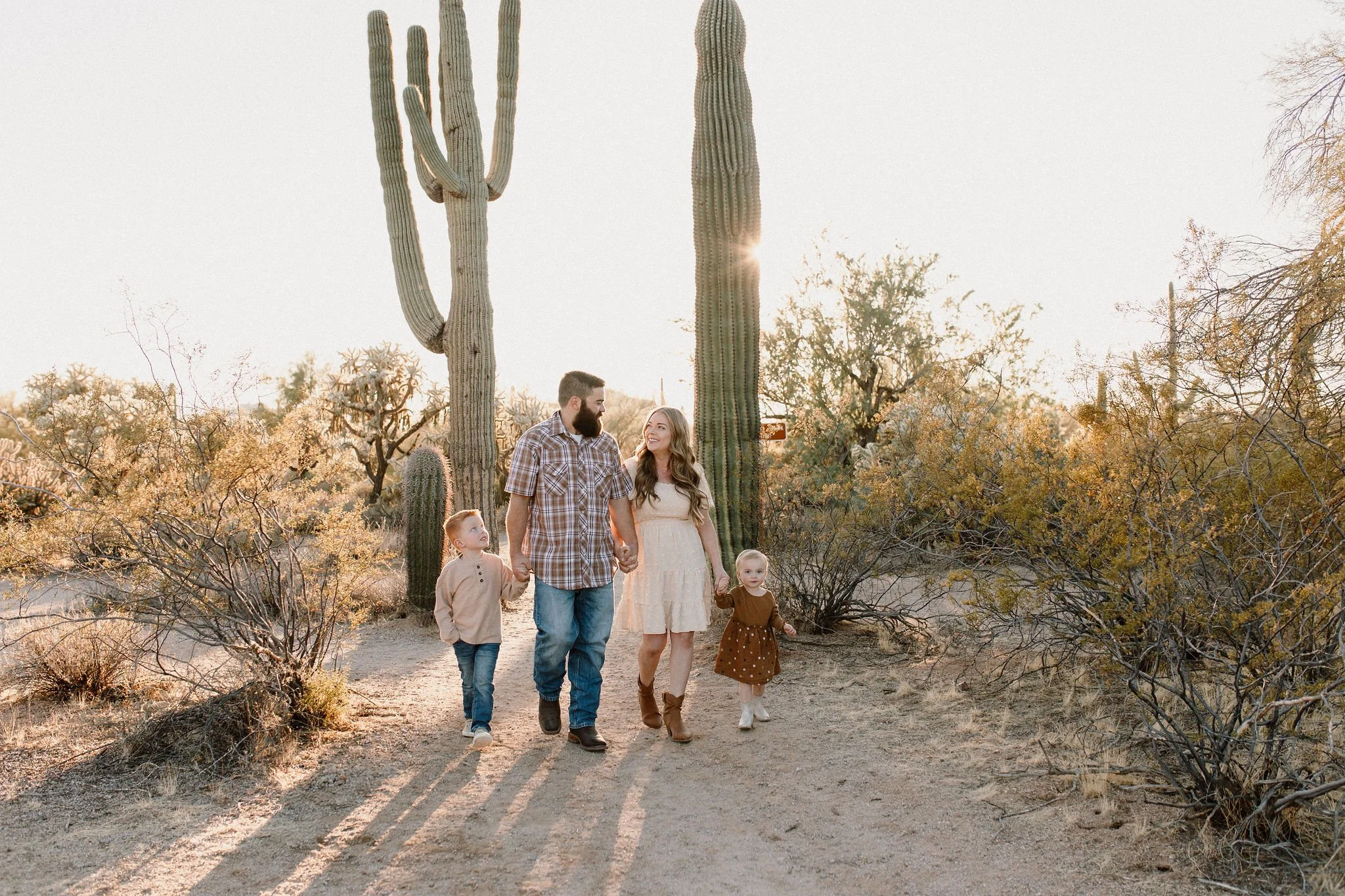 family of four holding hands and walking in desert with large saguaros during family photo session at Usery Mountain in Arizona