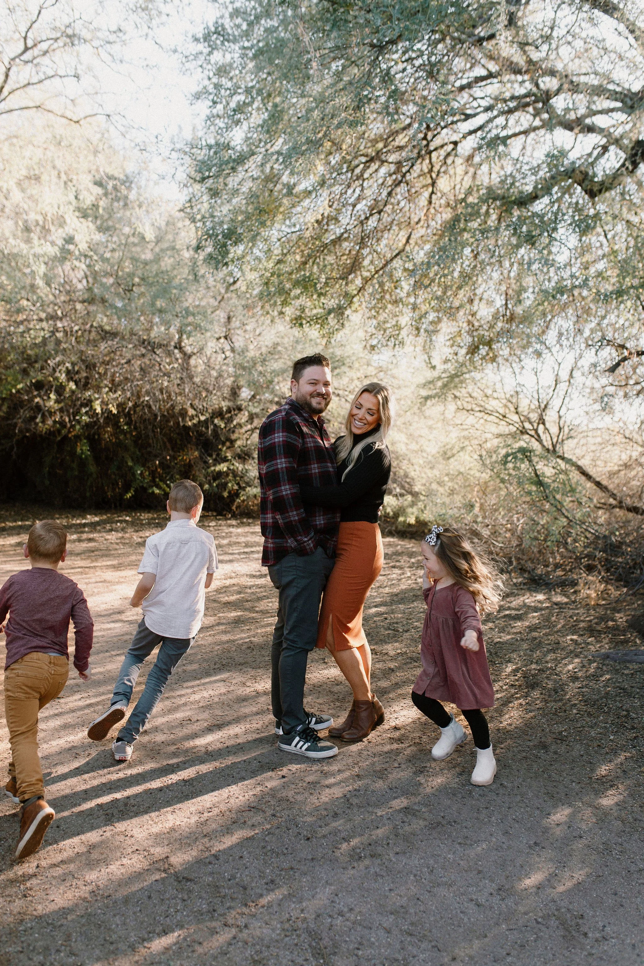 mom and dad hugging while their three children run around them at Gilbert Riparian Preserve in Arizona during family photo session